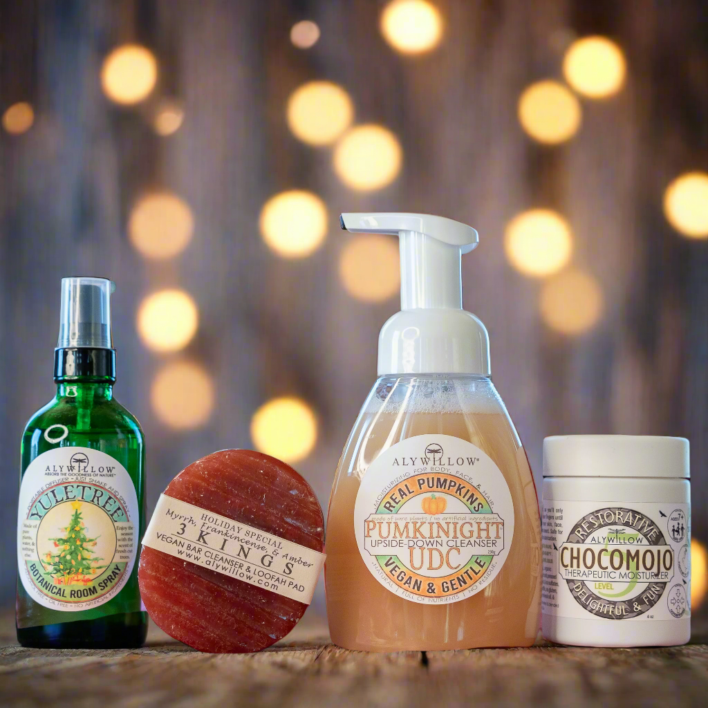 A collection of Aly Willow products including a pumpkin-scented foaming cleanser, a bar of soap, a room spray, and a moisturizer, all arranged on a wooden surface with Christmas tree lights in the background.