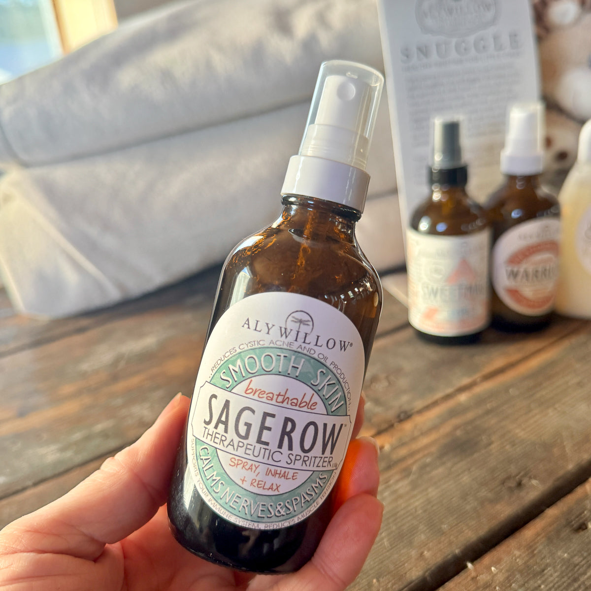 Bottle of Alywillow Sagerow Smooth Skin Therapeutic Spritzer held by a hand on a wooden surface with other products in the background.