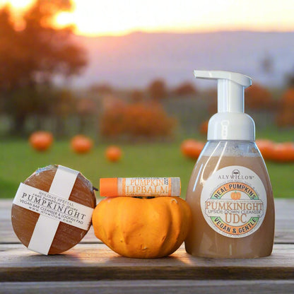 The image shows a Pumpkin Spice Gift Set which includes a pumpkin-scented lip balm, a pumpkin-scented bar cleanser, and a pumpkin-scented UDC cleanser, all branded as 'Aly Willow'. The products are displayed on a wooden surface with a backdrop of pumpkins and a sunset.