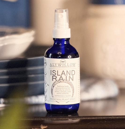 A cobalt blue glass spray bottle of Aly Willow Island Rain Medicinal Plant Spray with a white spray cap and white label, sitting on a dark wooden surface with a blurred background.