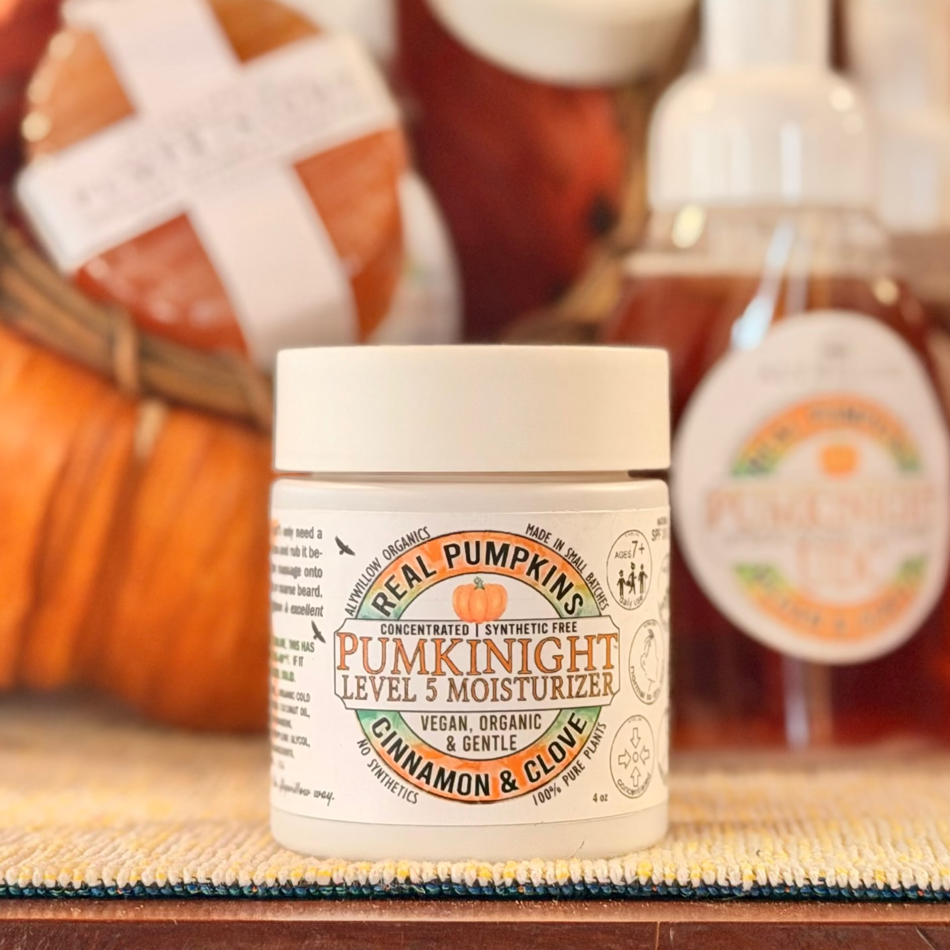 A jar of Real Pumpkins Pumpkinight Level 3 Moisturizer in Cinnamon & Cloves sits on a woven mat in front of a blurred background of pumpkins and other skincare bottles.