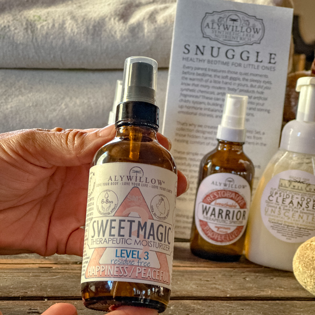 Hand holding a bottle of AlyWillow Sweet magic Level 3moisturizer with other products in the background.